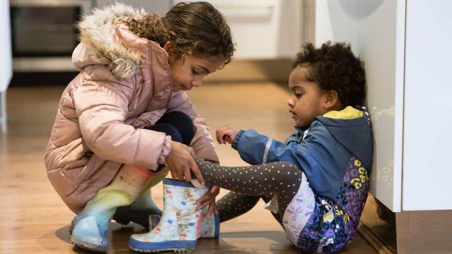 Sisters, dressed in their warm coats, getting ready to go outside and play. The older sibling is helping the younger sister put on her wellington boots.