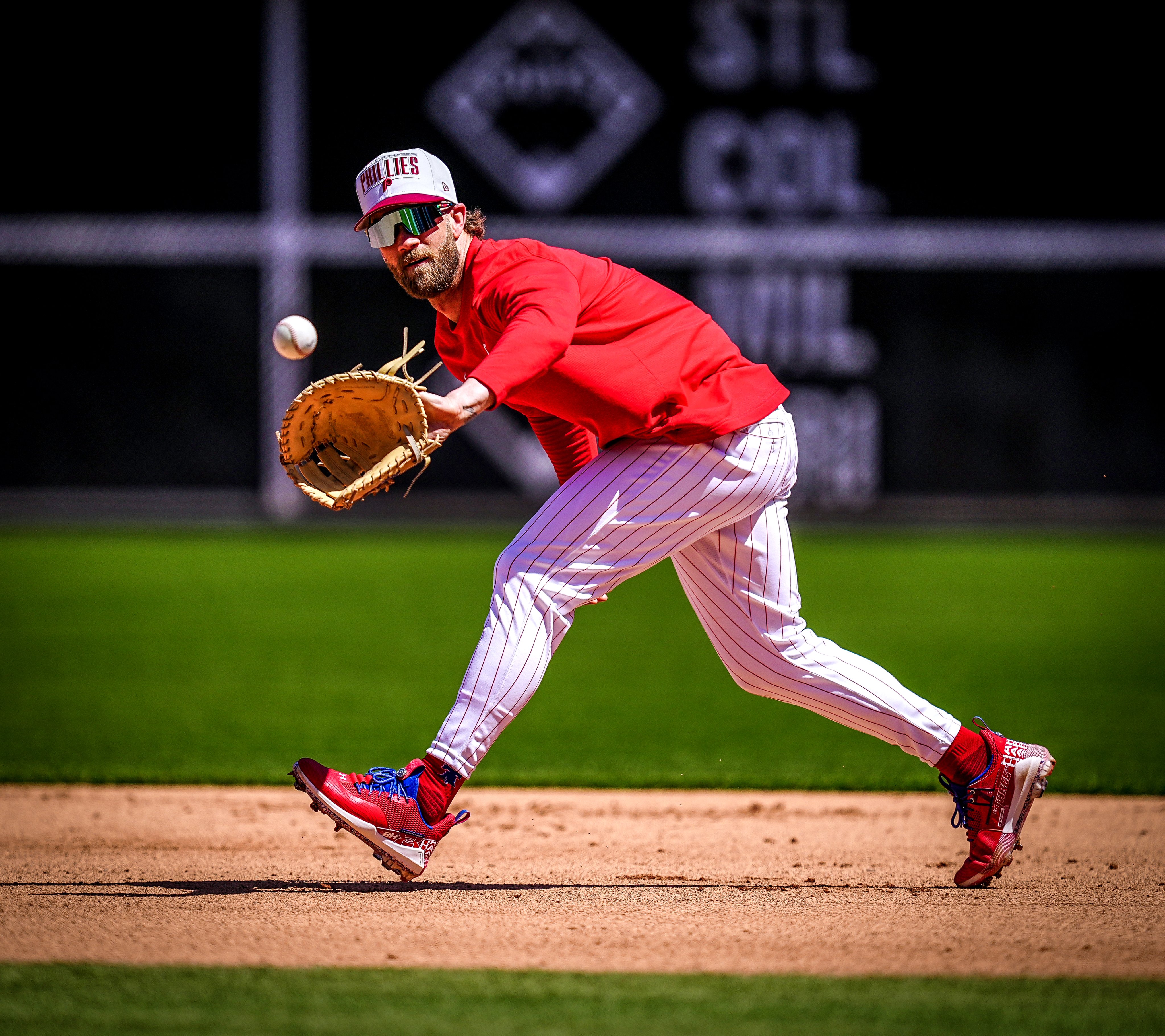 Bryce Harper about to catch a ball during his on-field session at Citizens Bank Park. He's wearing a red long-sleeve Phillies shirt, red pinstripes pants, and a white hat.